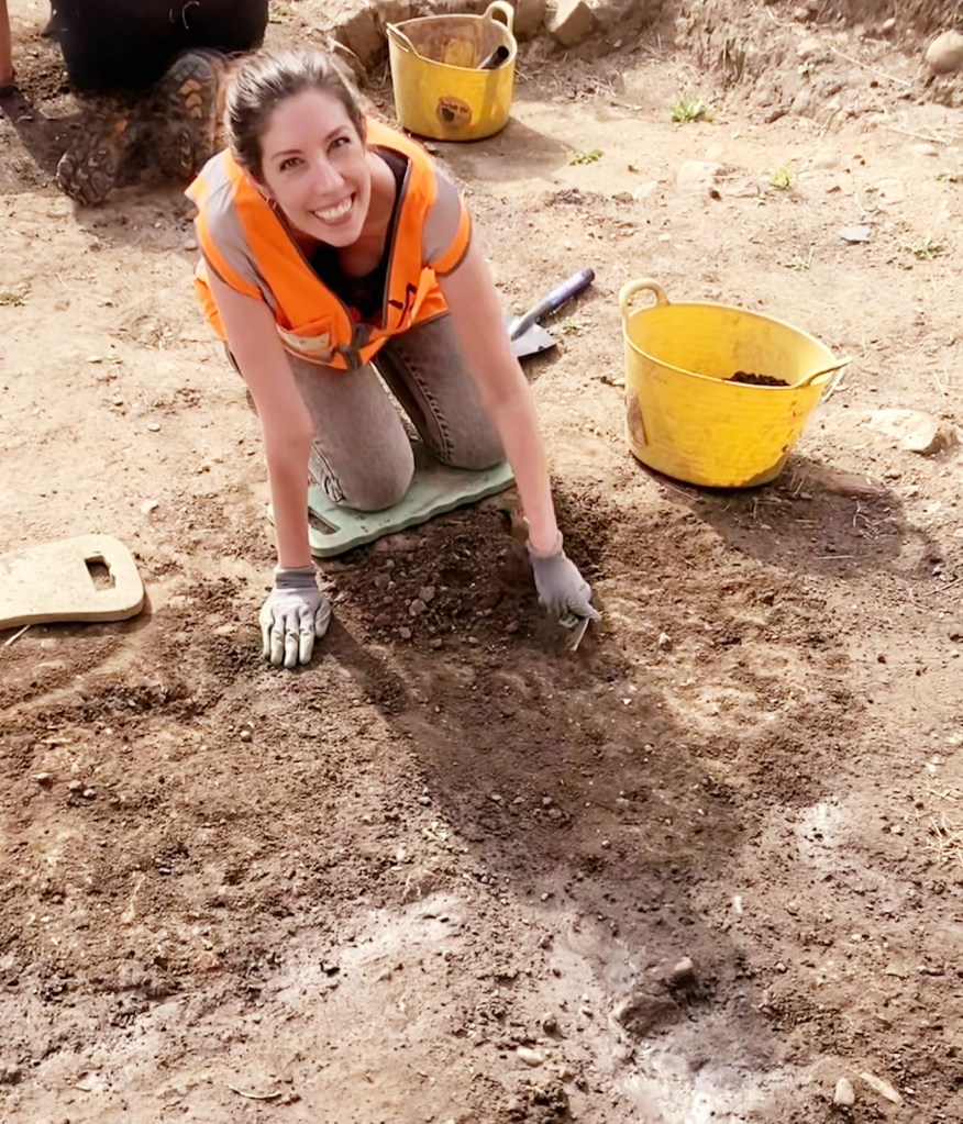 Image of Emily Peacock. Woman smiling at camera while digging archaeology.